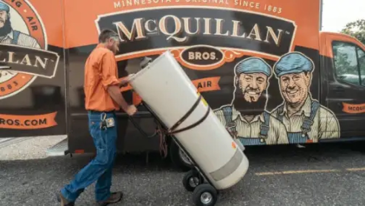 Worker moving a wrapped water heater on a dolly past a McQuillan Bros branded van