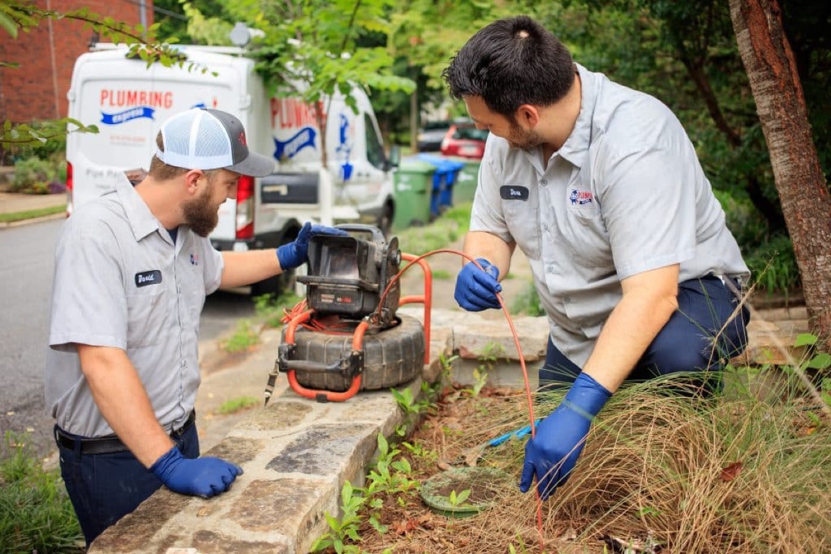 Two technicians operating a drain cleaning machine at a curb with branded vans behind