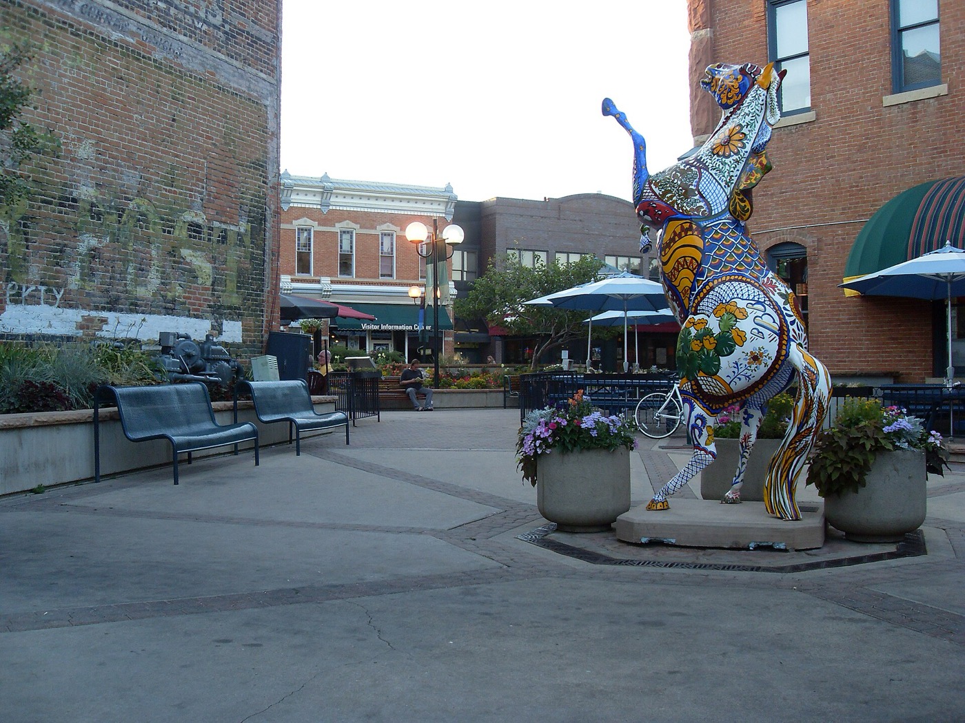 A painted horse sculpture and café umbrellas on a plaza in Old Town Fort Collins, with historic brick buildings behind