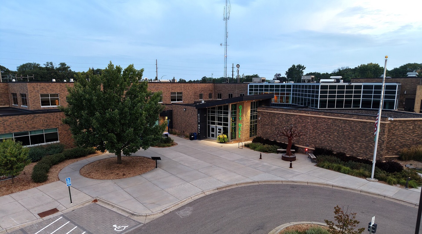Brick Eagan City Hall entrance with a lit green sign, plaza and tree