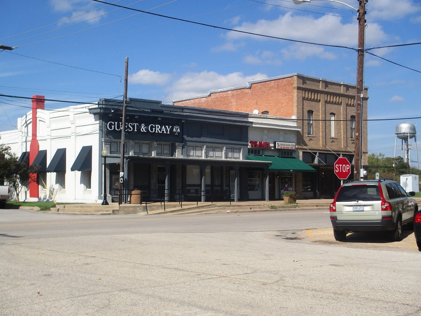 Brick and painted storefronts on a downtown street corner in Forney, Texas, with a water tower in the distance